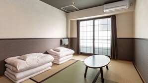 Habitación de hotel con esteras de tatami, una mesa de tatami, un sofá con cojines, una puerta corredera de vidrio y un equipo de aire acondicionado.