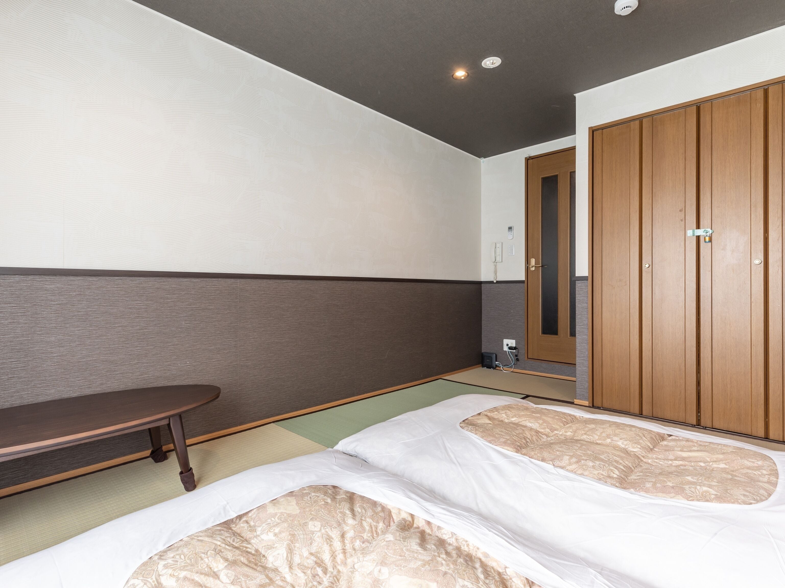 A hotel room with a bed, a wooden wardrobe, a small table, and a wall-mounted light fixture.