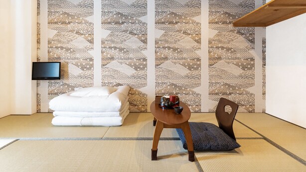 Una habitación japonesa tradicional con suelo de tatami, una mesa de madera baja con un juego de té, una cama futón y un televisor montado en la pared.