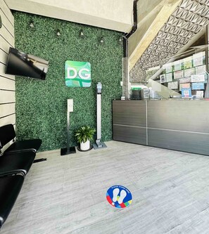 Un área de recepción con una pared verde, un letrero con el logotipo DG, una planta en maceta y un cartel que dice 'Por favor, pare aquí'.