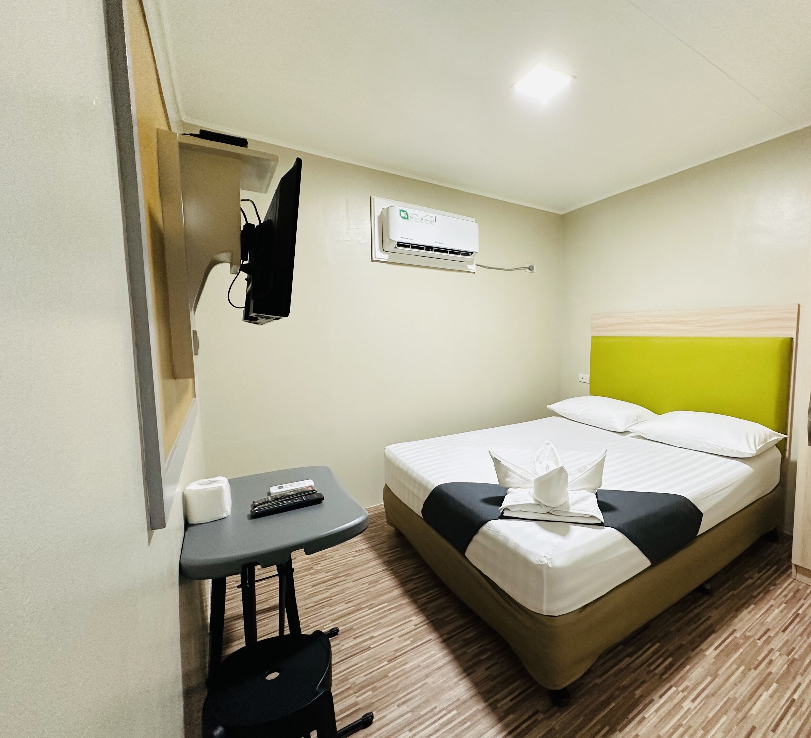 A hotel room with a bed, a small table with a remote control, a chair, an air conditioning unit, and a wall-mounted TV.