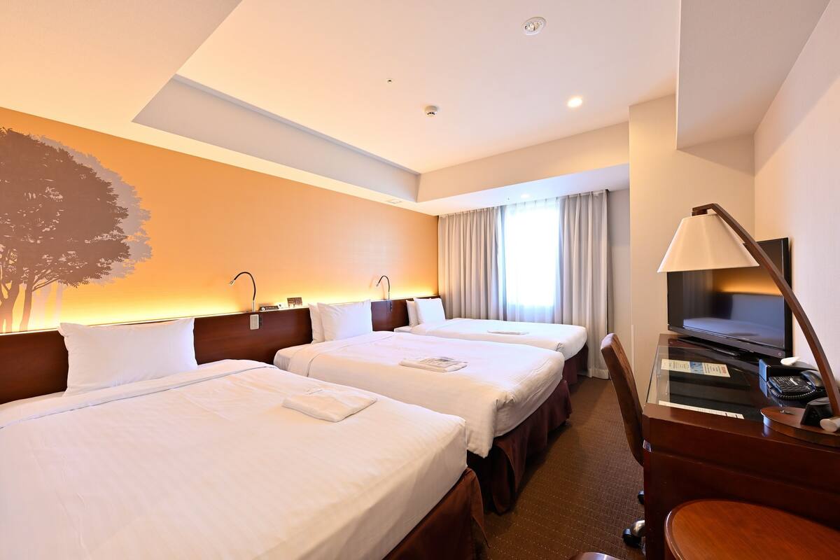 Habitación de hotel con dos camas, un escritorio con lámpara, televisor y un ventanal con cortinas.
