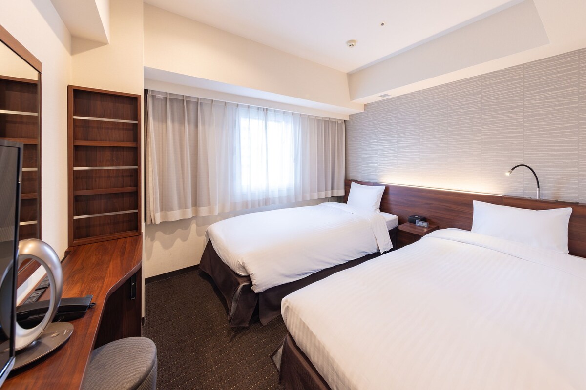 A hotel room with two beds, a desk with a chair, a TV, and a window with curtains.