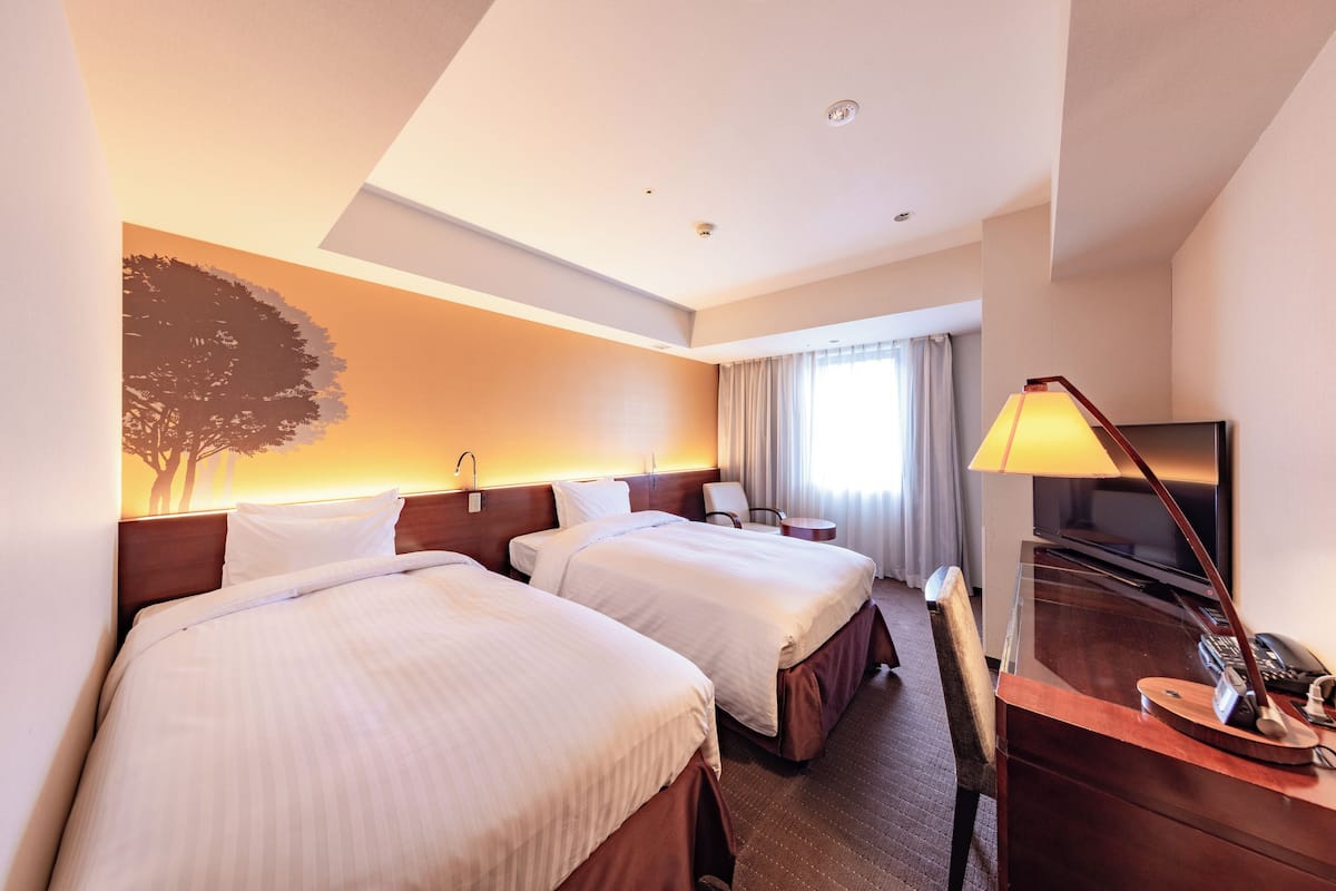 Habitación de hotel con dos camas, un escritorio con lámpara, televisor y ventana grande con cortinas.