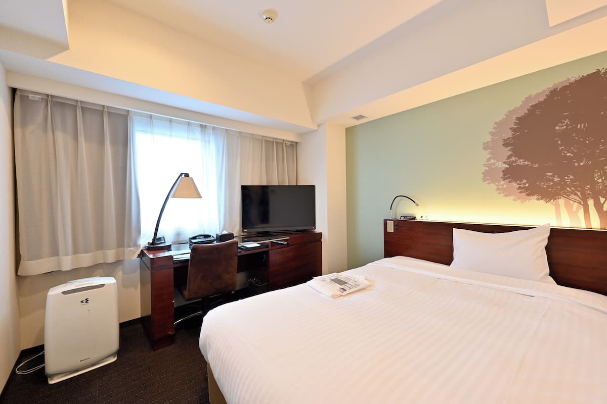 Una habitación de hotel con cama, escritorio, computadora, televisor y lámpara de pared.