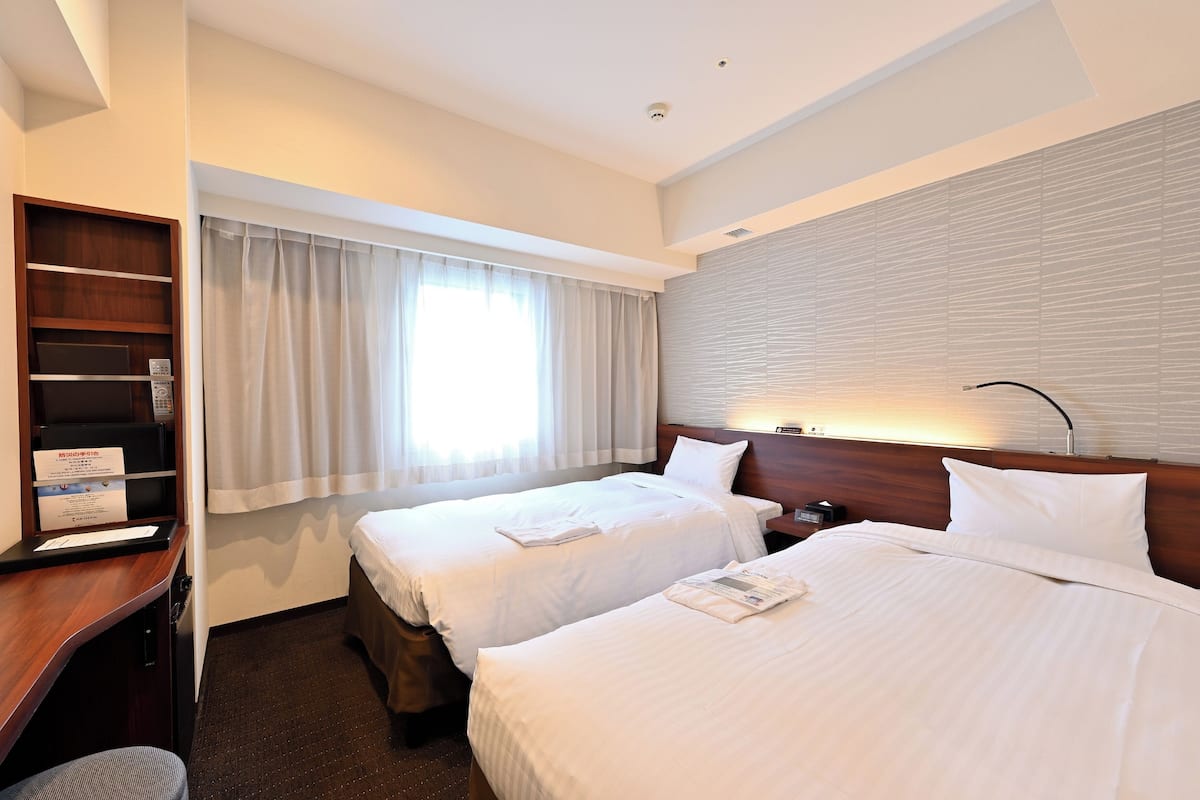 Habitación de hotel con dos camas, un escritorio, una estantería con objetos y una ventana con cortinas.