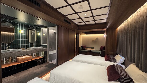Hotelkamer met twee bedden, een zithoek met een tafel, een badkamer met wastafel en een groot raam.