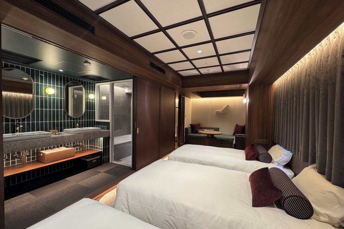 A hotel room with two beds, a sitting area with a table, a bathroom with a vanity, and a large window.