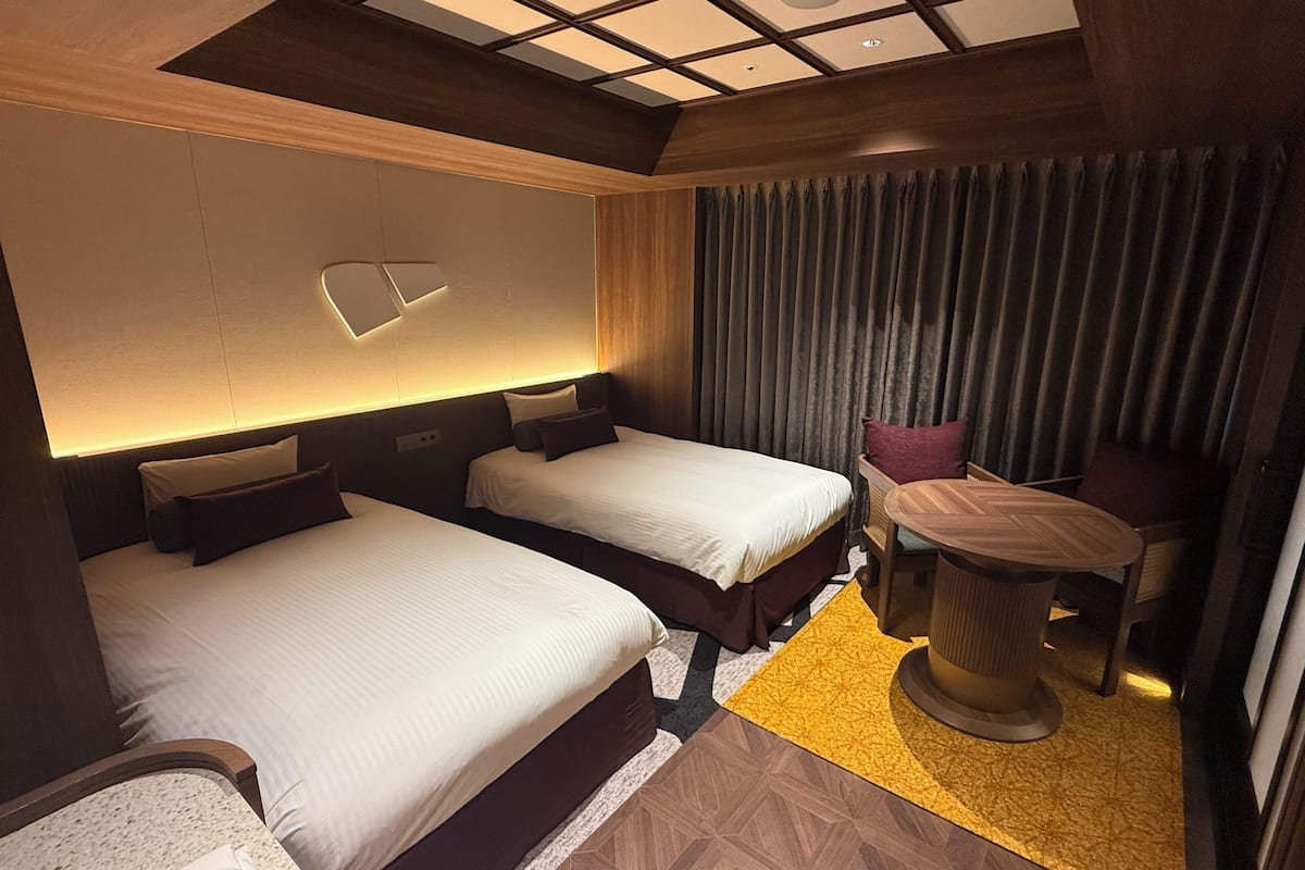 A hotel room with two beds, a small table, a chair, a wall-mounted light, and a curtain.