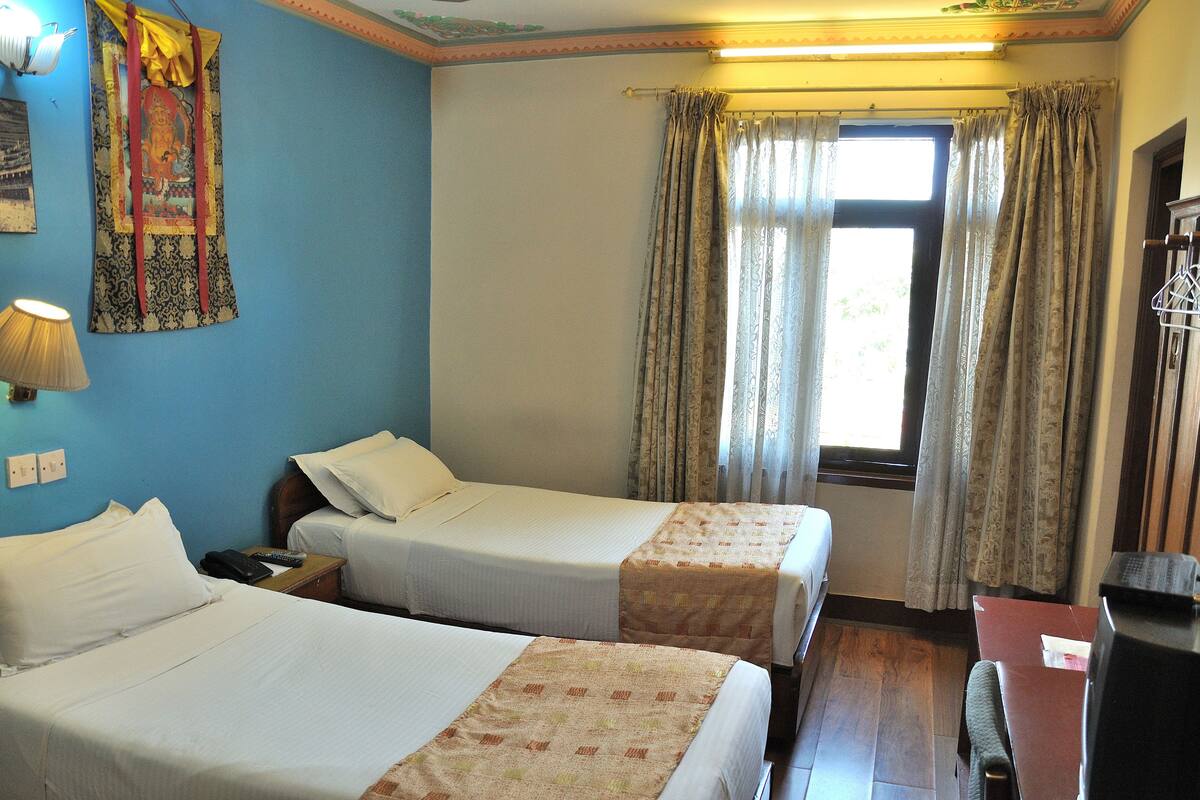Habitación de hotel con dos camas individuales, un escritorio, una silla, una lámpara, una ventana con cortinas y un tapiz en la pared.