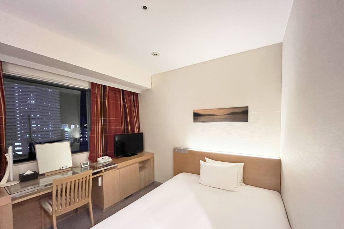 Ein Hotelzimmer mit einem Bett, einem Schreibtisch mit Computer, einem Fernseher und einem Fenster mit Vorhängen.
