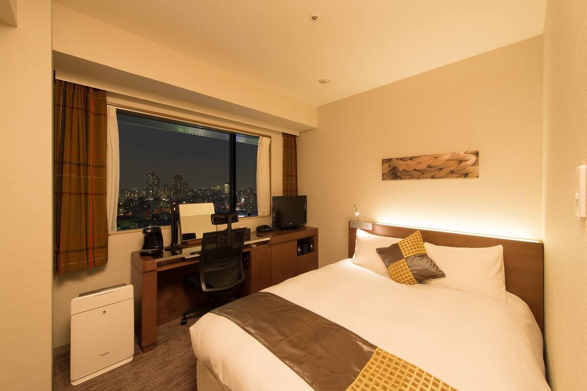 Ein Hotelzimmer mit einem großen Bett, einem Schreibtisch mit Computer, einem Stuhl und Blick auf die beleuchtete Stadt.