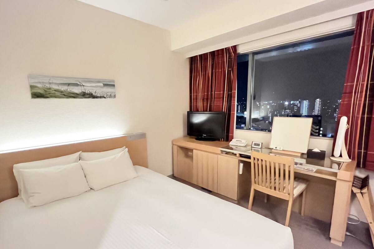 Ein Hotelzimmer mit einem großen Bett, einem Schreibtisch mit Computer, einem Fernseher und einem Fenster mit Blick auf die Stadt.