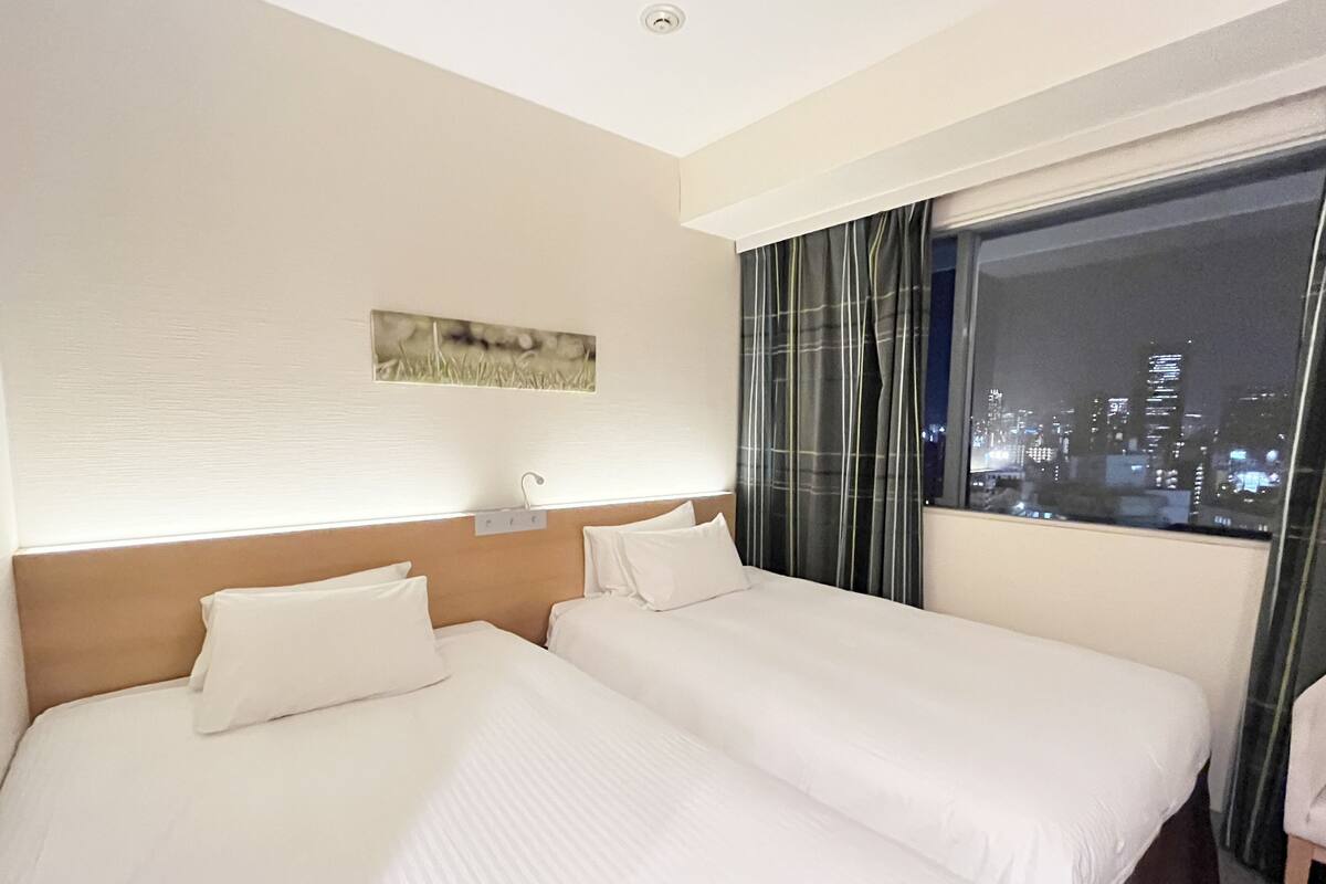 Ein Hotelzimmer mit zwei Betten, einem Nachttisch, einem Bild an der Wand und Blick auf die Stadt durch das Fenster.