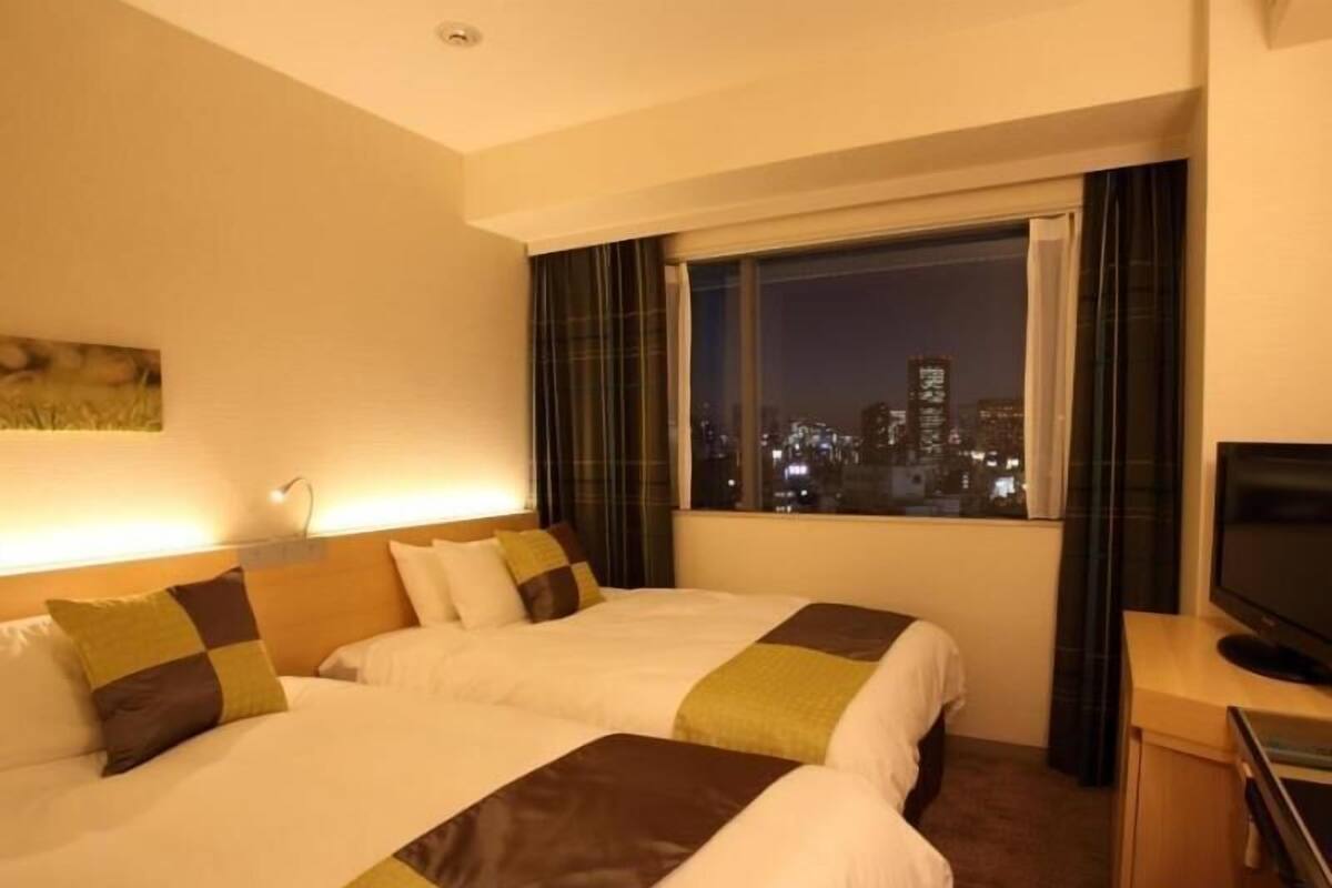 Ein Hotelzimmer mit zwei Betten, einem Fernseher, einem Fenster mit Blick auf die Stadt und einem Bild an der Wand.