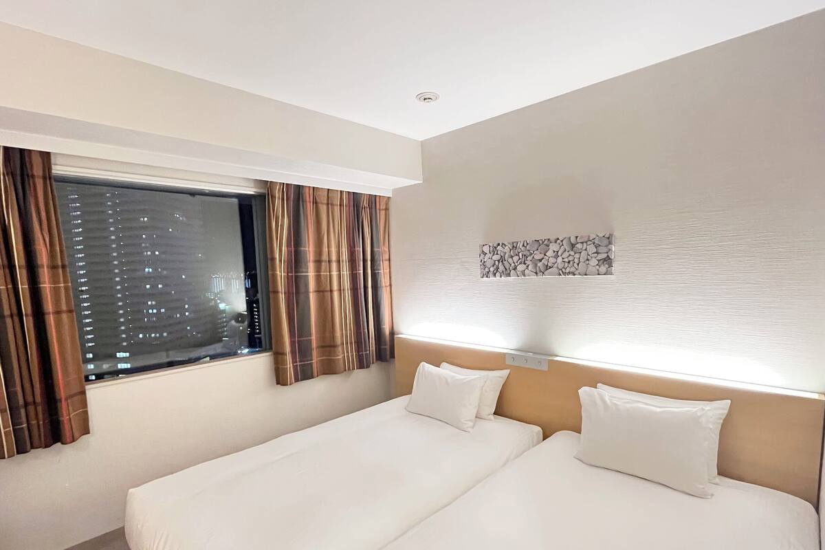 Ein Hotelzimmer mit zwei Einzelbetten, einem Fenster mit Vorhängen, einer Wandleuchte und einer Wanddekoration mit Struktur.