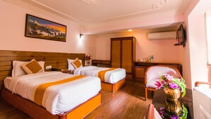 A hotel room with two beds, a wooden headboard, a painting, an air conditioning unit, a desk with a chair, and a vase with flowers. - Hotel Blue Horizon (Kathmandu)