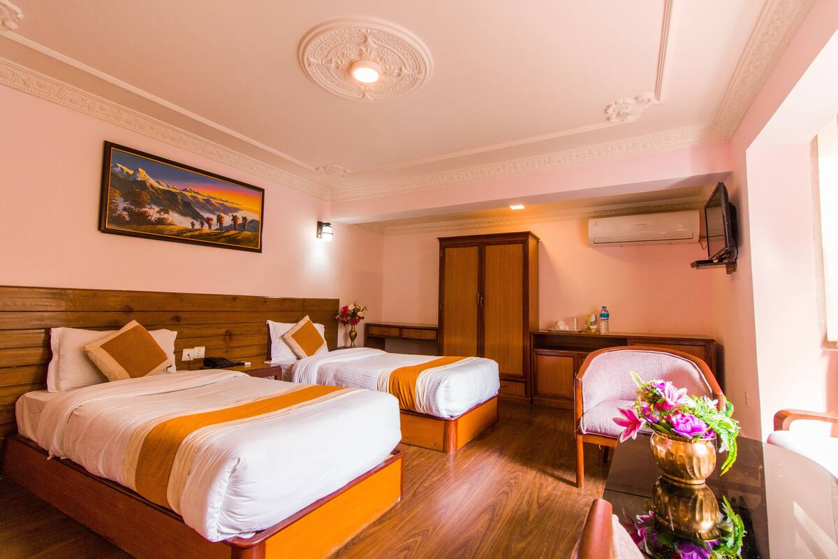 A hotel room with two beds, a wooden headboard, a painting, an air conditioning unit, a desk with a chair, and a vase with flowers.