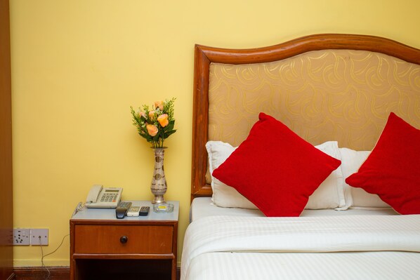 A neatly made bed with red pillows, a bedside table with a phone, a vase with flowers, and a wall socket. - Hotel Blue Horizon (Kathmandu)