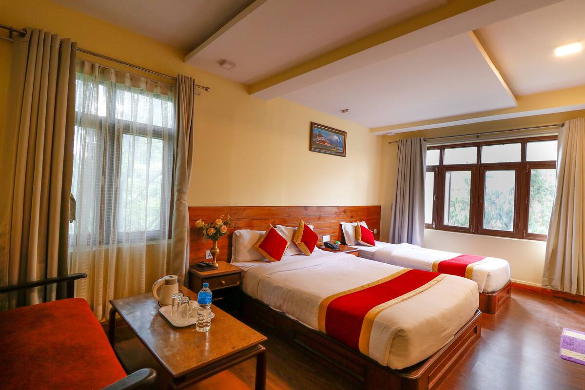 A hotel room with two beds, a wooden headboard, a table with a kettle and water bottle, and a window with curtains.