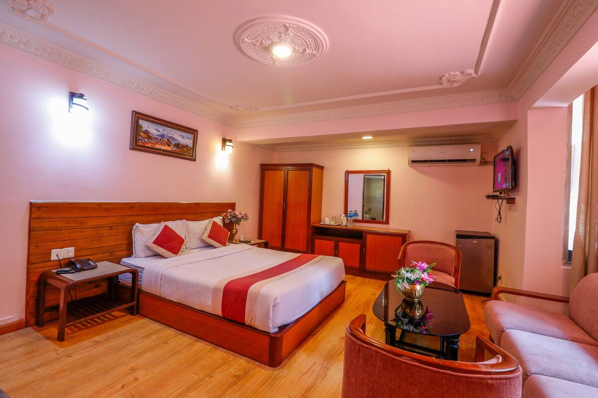 A hotel room with a large bed, a desk, a chair, a small table with flowers, a TV, and a wardrobe.