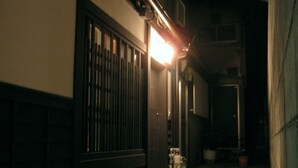 A narrow alleyway with a building on one side and a wall on the other. There is a light source at the end of the alleyway. - Kagariya (Kyoto)