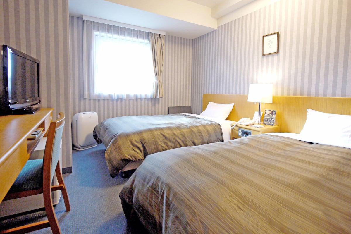 A hotel room with two beds, a desk with a television, a bedside table with a lamp, and a window with curtains.