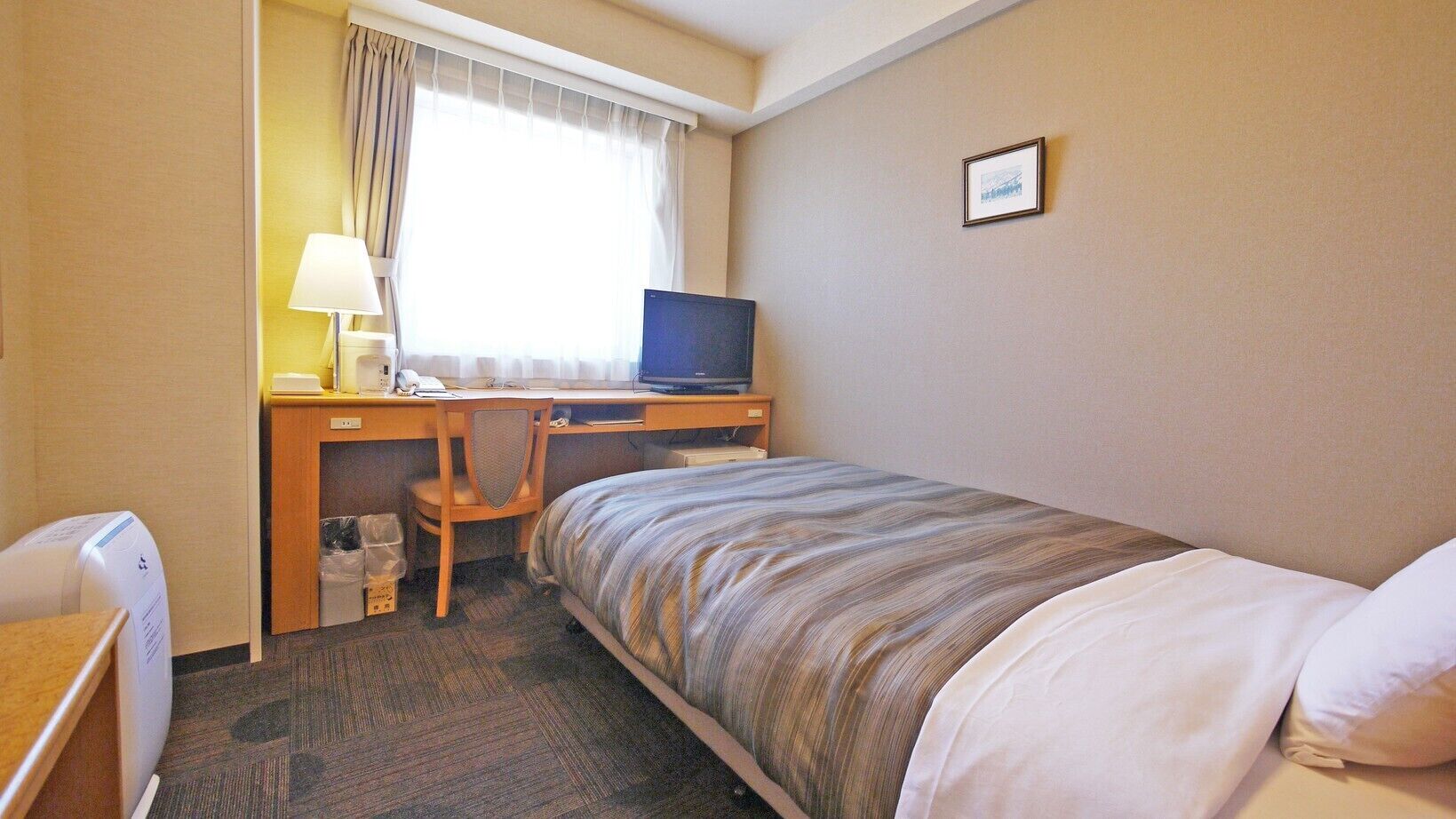 A hotel room with a bed, a desk with a computer, a chair, a lamp, and a small trash bin.