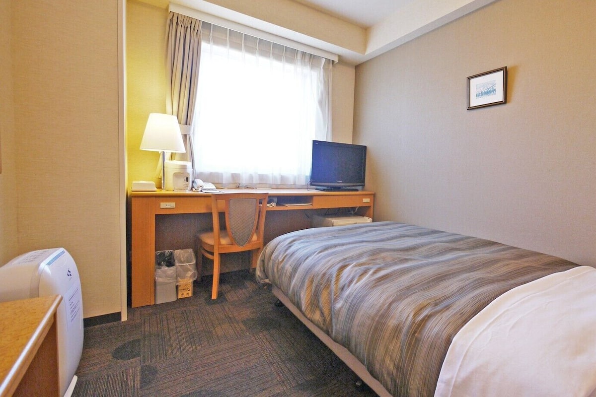 A hotel room with a bed, a desk with a computer, a chair, a lamp, and a small trash bin.
