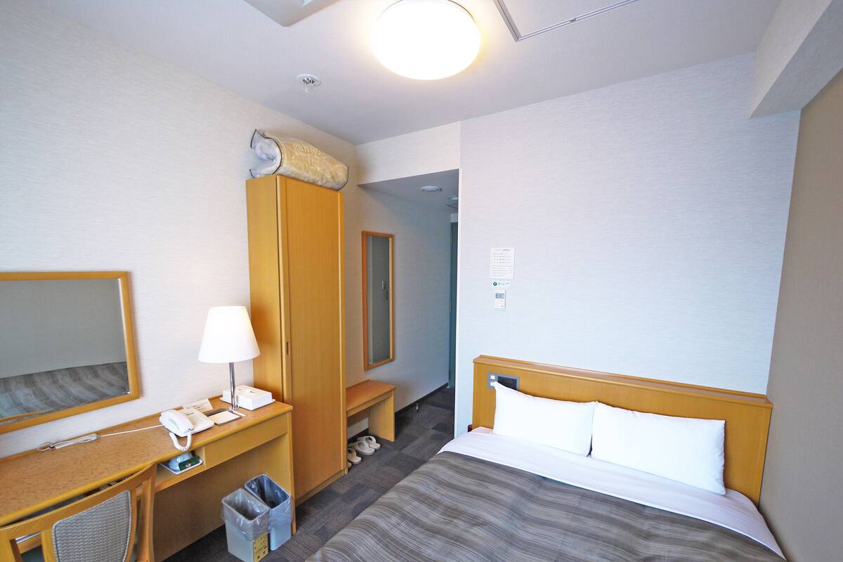 A hotel room with a bed, a desk with a telephone and lamp, a mirror, and a trash bin.