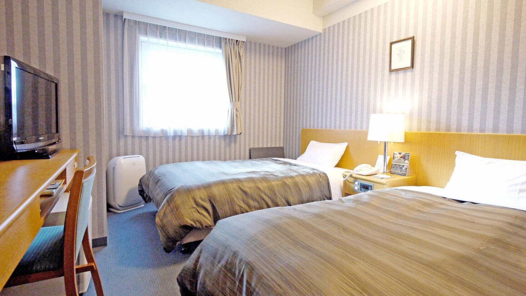 A hotel room with two beds, a desk with a television, a bedside table with a lamp, and a window with curtains.