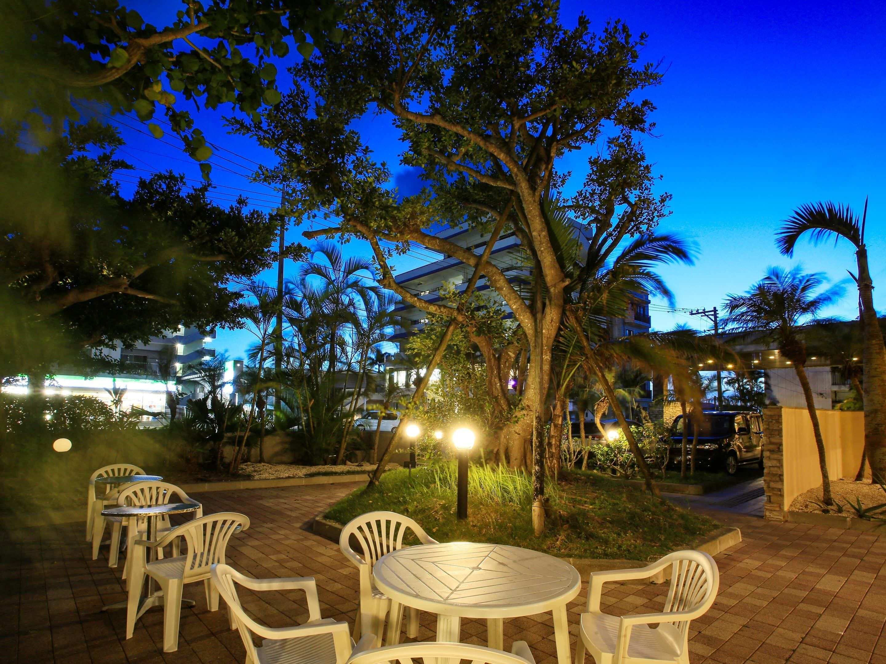An outdoor seating area with white plastic chairs and tables, surrounded by trees and a building in the background.