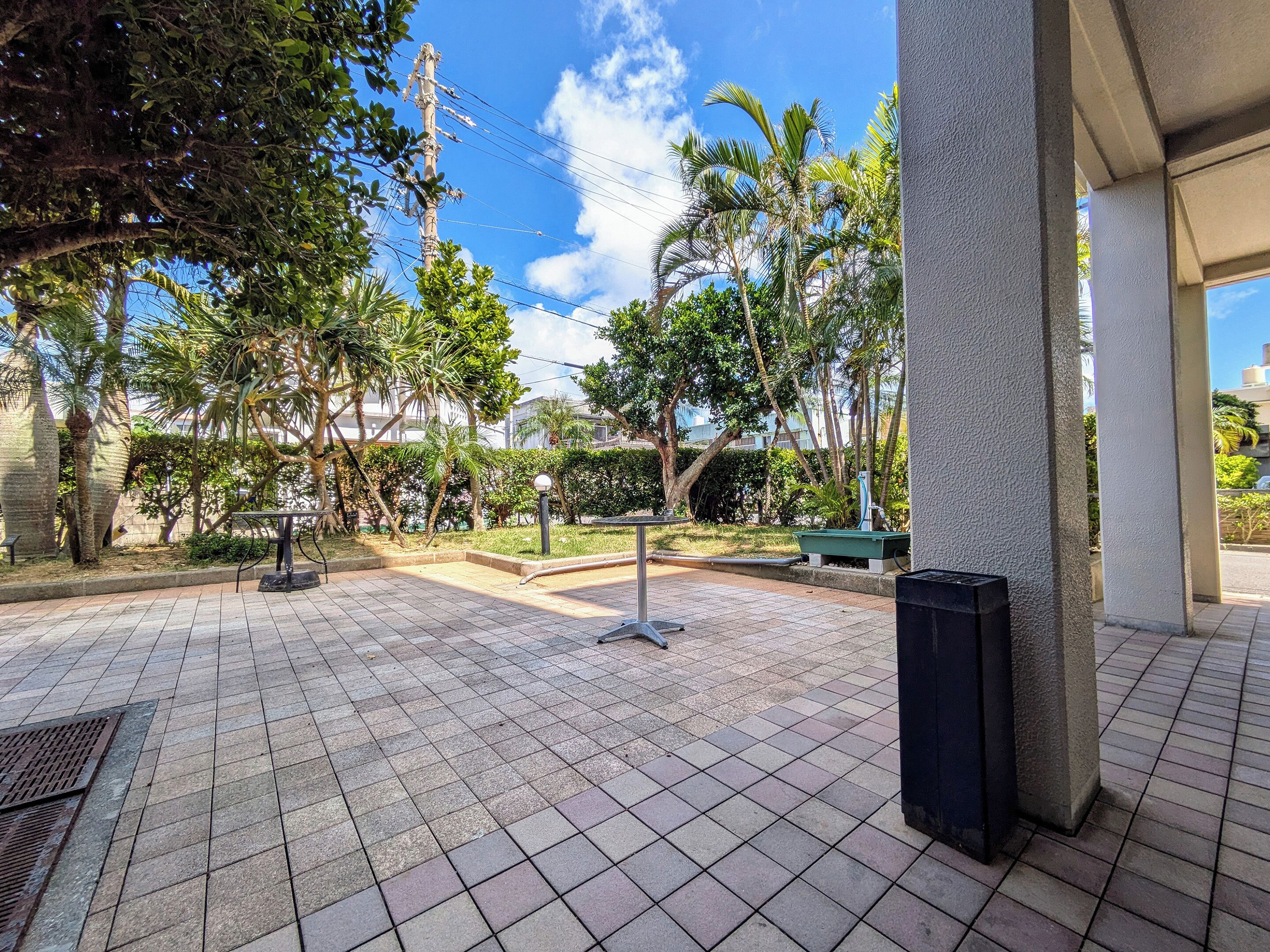 A paved outdoor area with tiled flooring, a metal bench, and a trash bin.
