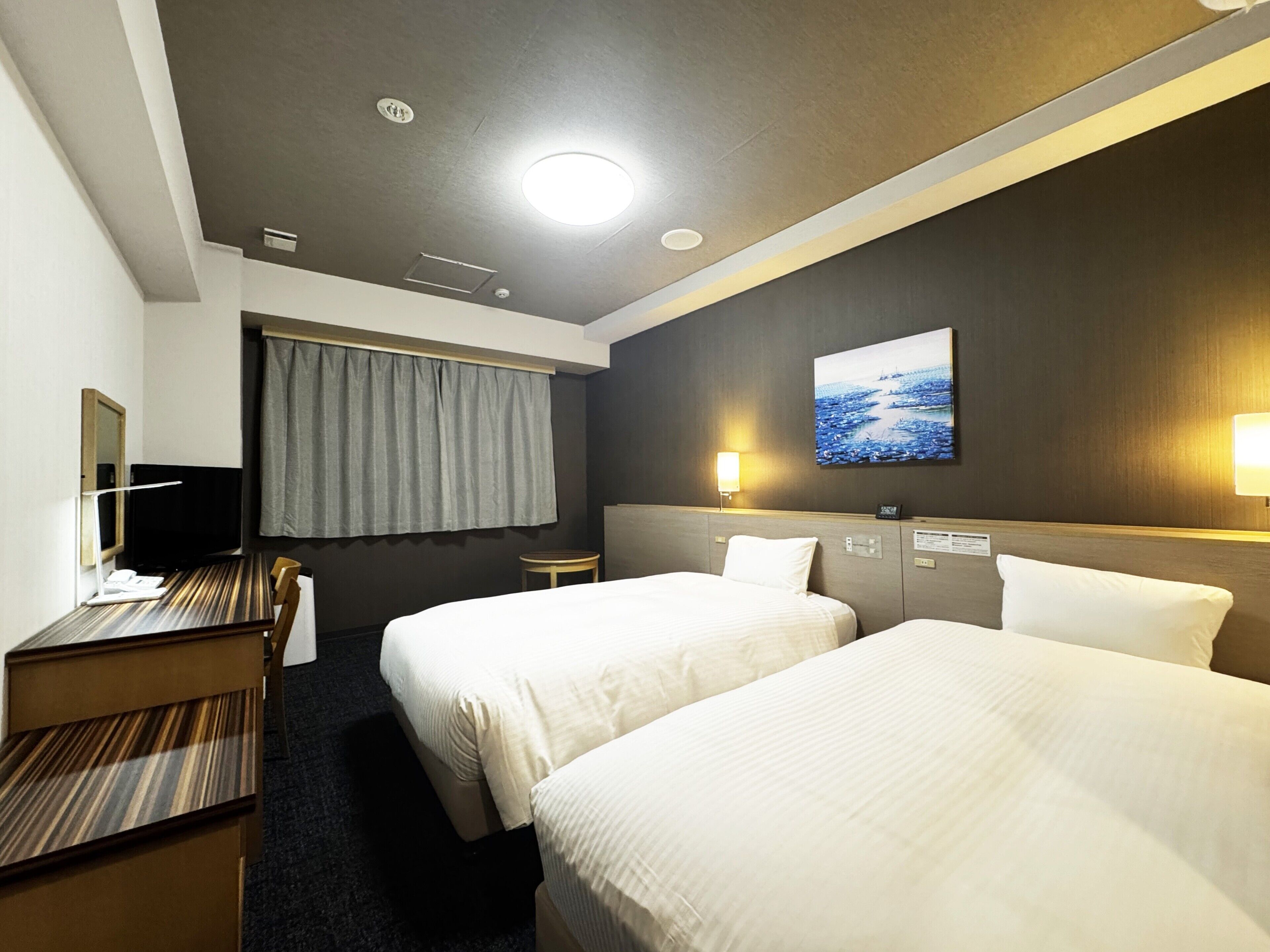A hotel room with two beds, a desk, a chair, a television, a window with curtains, and a painting on the wall.