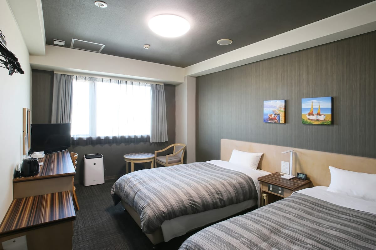 Ein Hotelzimmer mit zwei Betten, einem Schreibtisch, einem Flachbildfernseher, einem kleinen Tisch und zwei Bildern an der Wand.