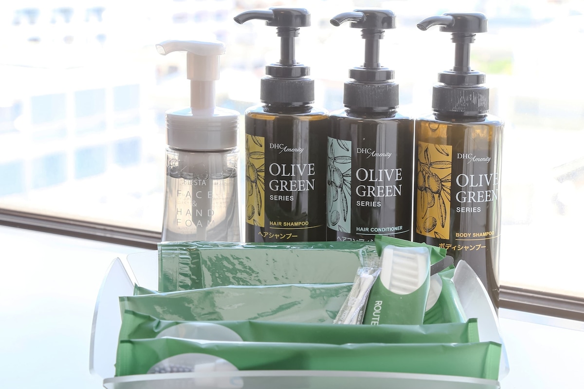 A bathroom with a window view, a green toiletry bag, and four bottles of DHc Amenity Olive Green Series hair products.