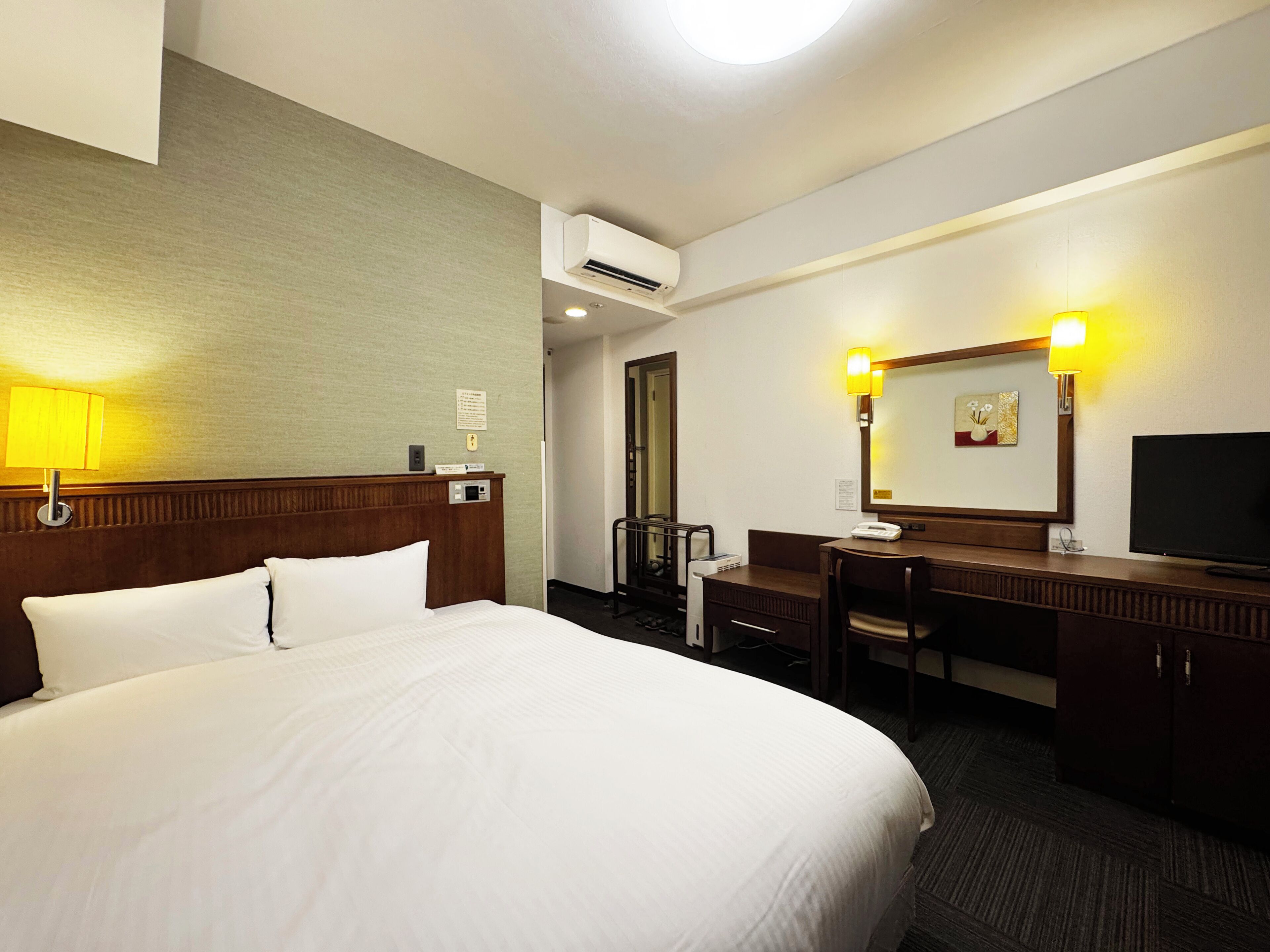 A hotel room with a large bed, a desk, a chair, a television, and an air conditioning unit.