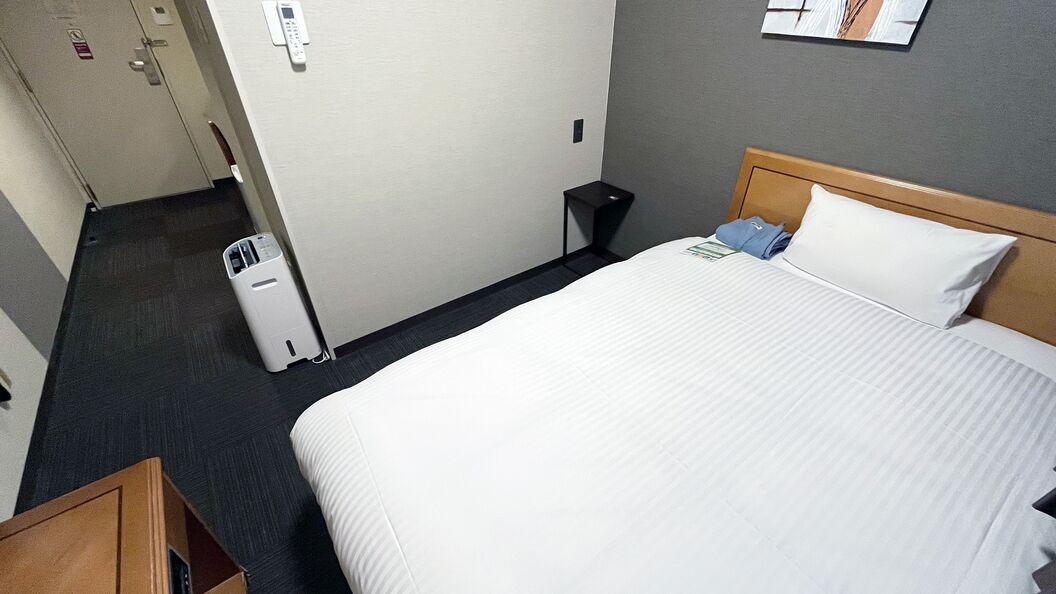 A hotel room with a bed, a small bedside table, a trash bin, and a wall-mounted control panel.