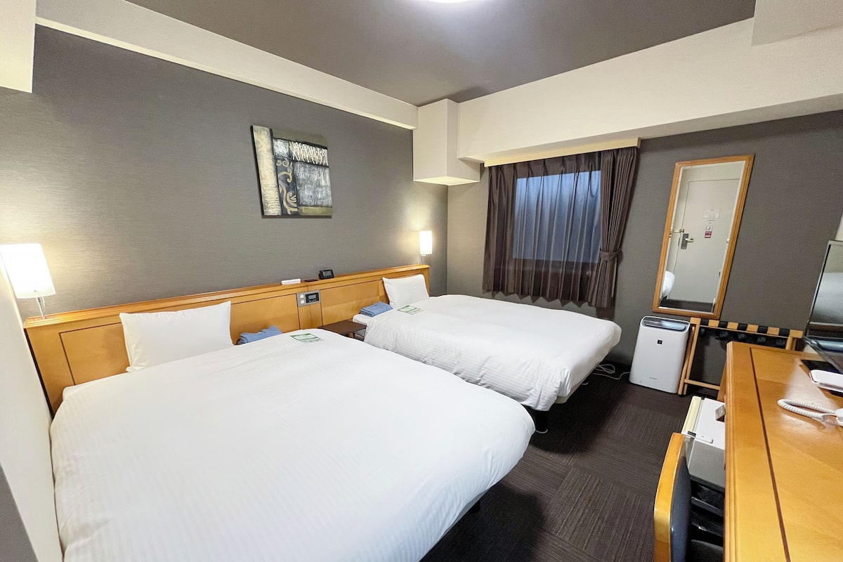 Une chambre d’hôtel avec deux lits, un bureau, une chaise, une lampe, un miroir et une fenêtre avec des rideaux.