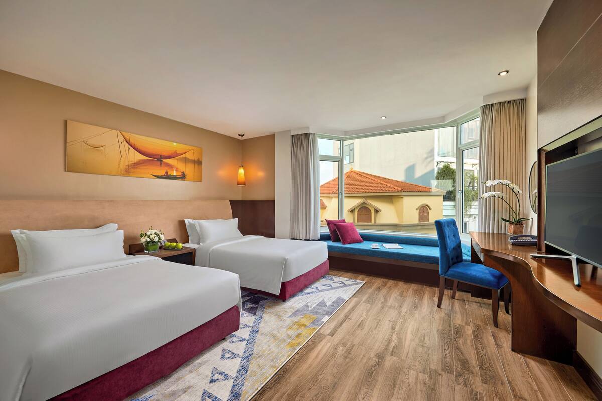 A hotel room with two beds, a desk with a TV, a window with a view of buildings, and a painting on the wall.