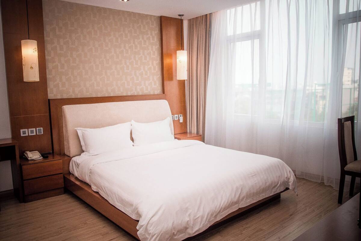 A hotel room with a large bed, wooden headboard, bedside lamps, a telephone, and a window with sheer curtains.