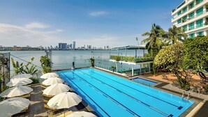 Una piscina con lettini e ombrelloni, con vista su uno specchio d'acqua e sullo skyline di una città sullo sfondo.