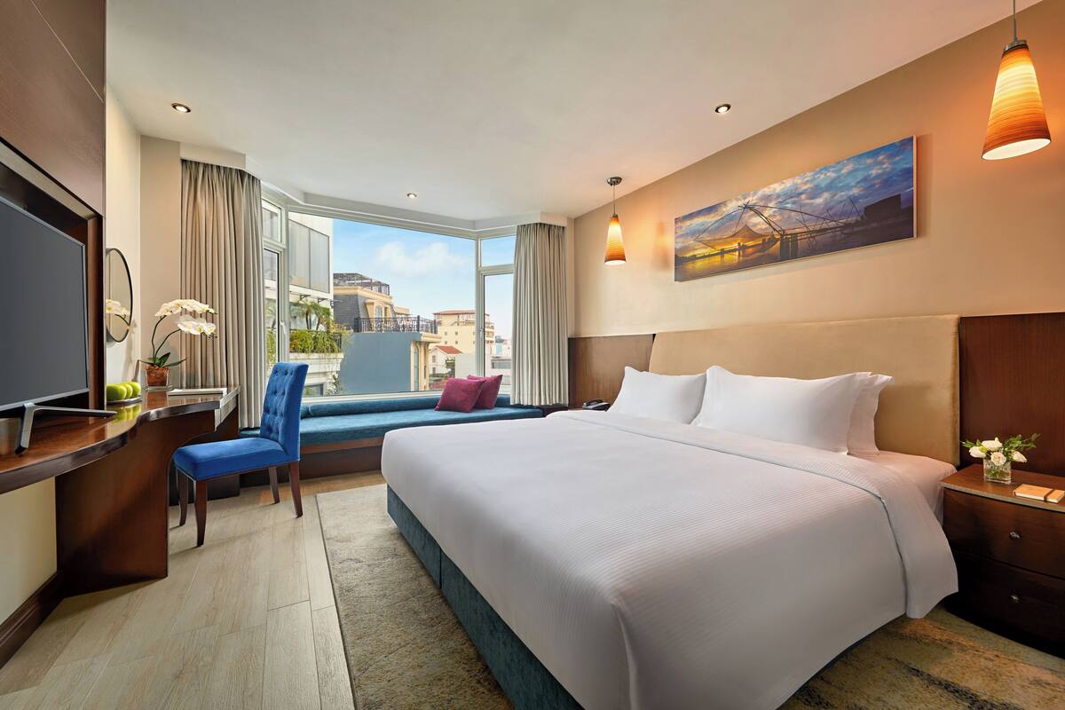 A hotel room with a large bed, a desk with a computer, a chair, a window with curtains, and a painting on the wall.