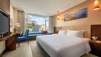 A hotel room with a large bed, a desk with a computer, a chair, a window with curtains, and a painting on the wall.