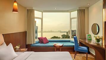 A hotel room with a large bed, a desk, a chair, a window seat with a view of a body of water, and a hanging light fixture.