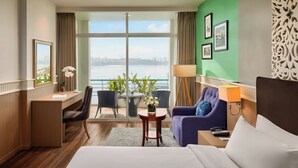 Camera d'albergo con scrivania, due poltrone, un divano, un letto, vista sull'acqua e un balcone con piante.