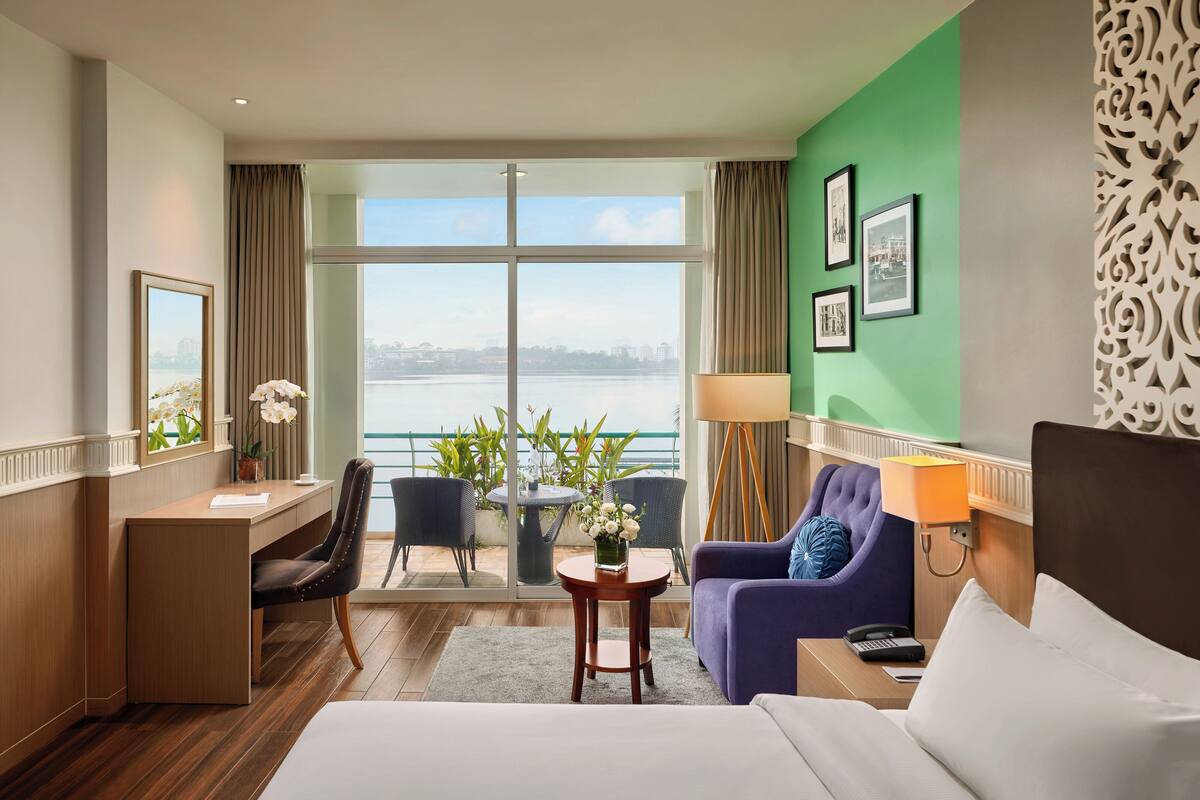 A hotel room with a desk, two chairs, a sofa, a bed, a view of the water, and a balcony with plants.