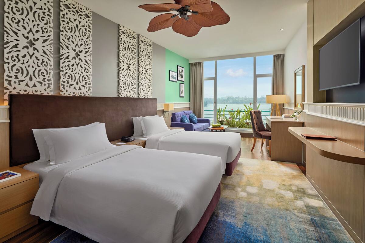 A hotel room with two beds, a desk, a chair, a TV, and a balcony with a view.