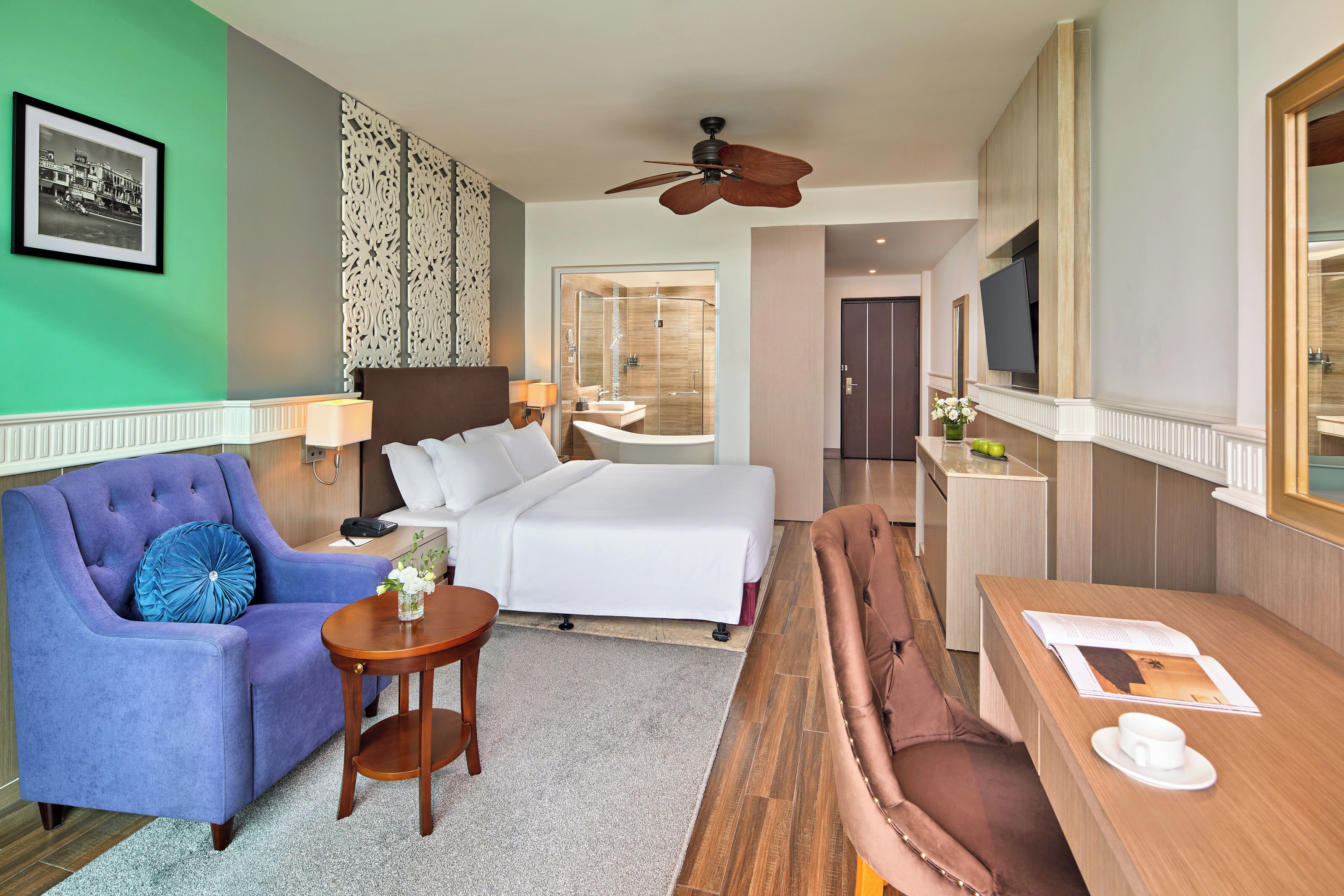 Habitación de hotel con cama, sofá, una mesita, televisor y baño visible a través de una puerta de cristal.