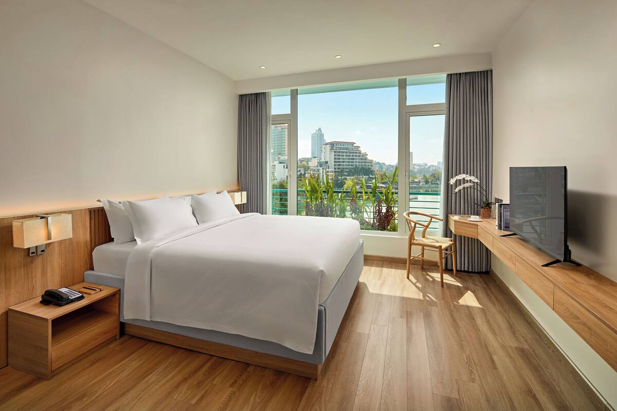 A modern hotel room with a large bed, a desk with a TV, and a view of the city through large windows.
